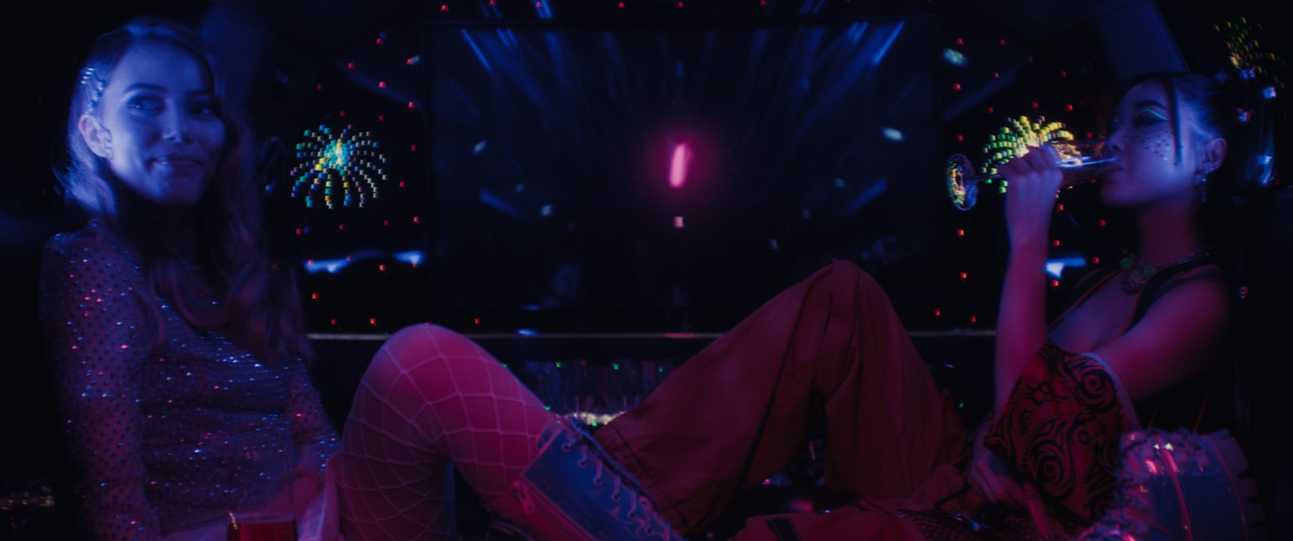 Two women sit in a dark, neon-lit room with colorful light reflections; one drinks from a glass while the other smiles, both dressed in party outfits.