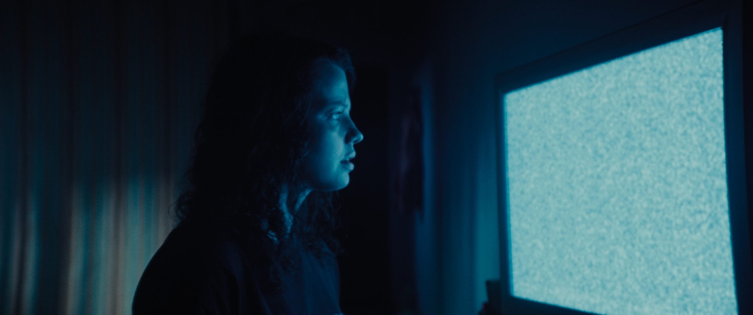 A person with curly hair looks at a television screen displaying static in a dimly lit room.