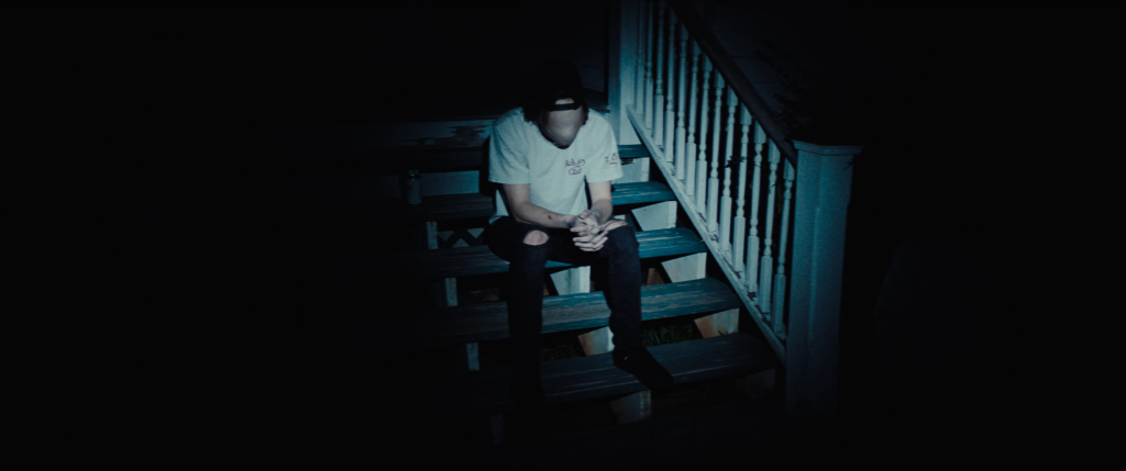 A person wearing a white t-shirt and cap sits alone on outdoor steps at night, with their face obscured by shadows and hands clasped.