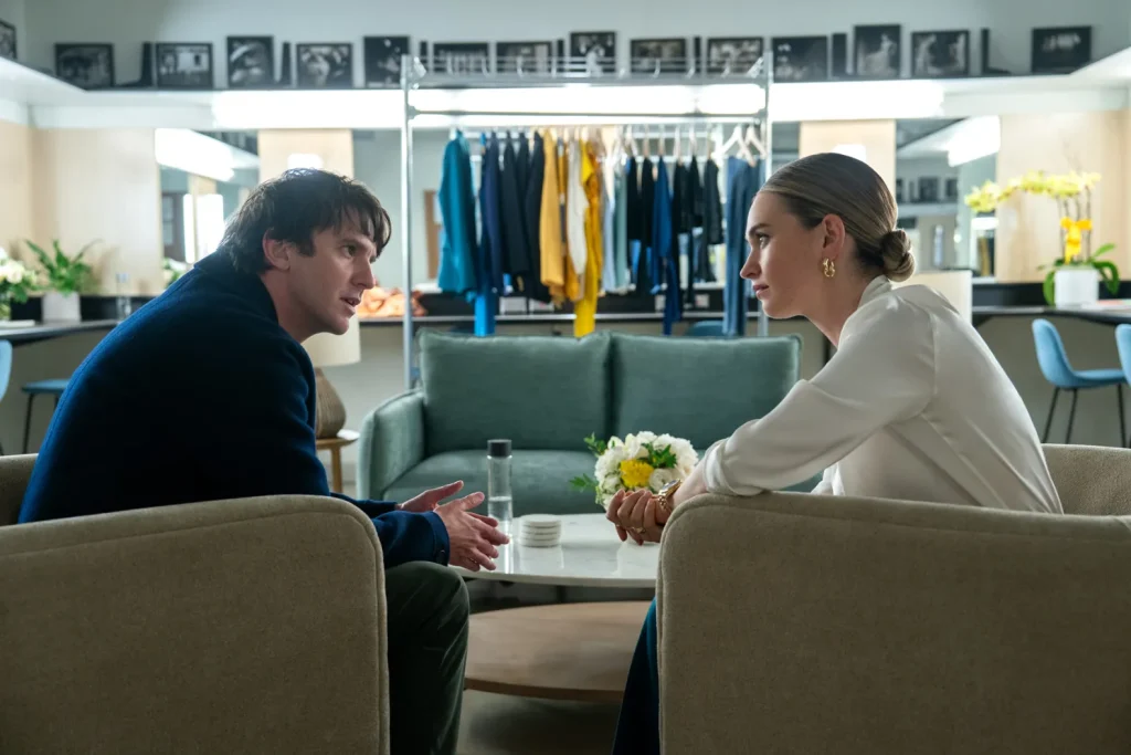 A man and a woman sit across from each other in a modern dressing room, engaged in a serious conversation, with clothing racks and a sofa in the background.