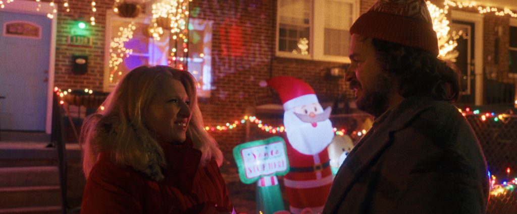 Two people stand outside at night, smiling at each other, with colorful Christmas lights and an inflatable Santa decoration in the background.