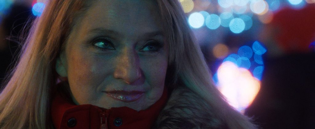 A woman with long blonde hair in a red coat looks to the side, smiling slightly, with colorful blurred lights in the background.