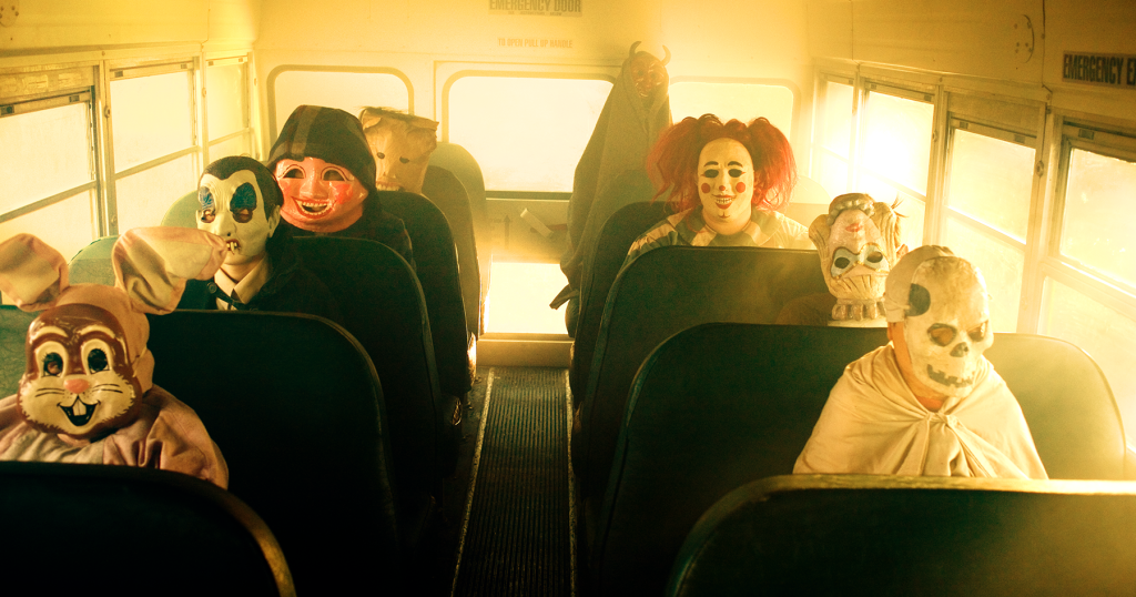 Children wearing various creepy costumes and masks sit quietly on a school bus with sunlight streaming through the windows.