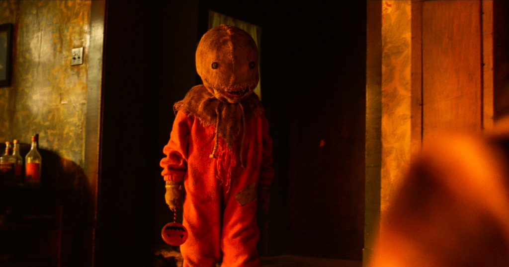 A person in an orange costume with a burlap sack mask stands indoors holding a small, round object, in a dimly lit room.