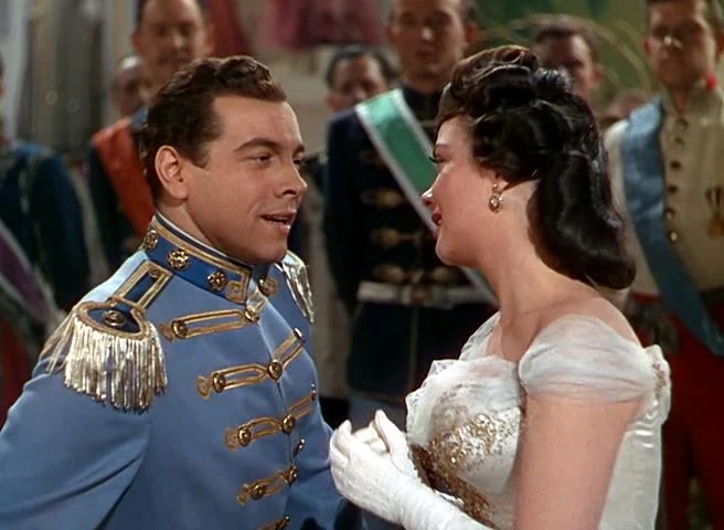 A man in a light blue military uniform speaks to a woman in a white gown; several people in formal attire are visible in the background.