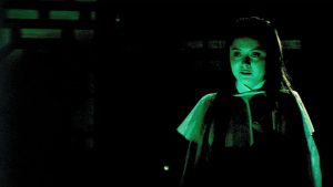 A girl with long dark hair stands alone in dim greenish light, looking forward with a neutral expression.