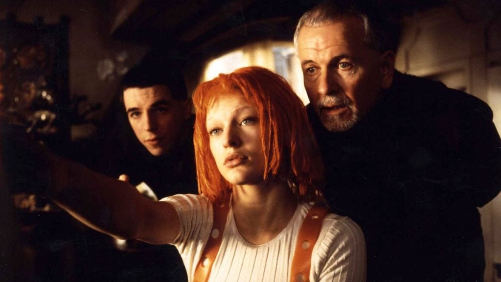 Three people indoors, with a woman with bright orange hair and two men looking in the same direction as she points offscreen.