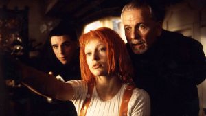 Three people indoors, with a woman with bright orange hair and two men looking in the same direction as she points offscreen.