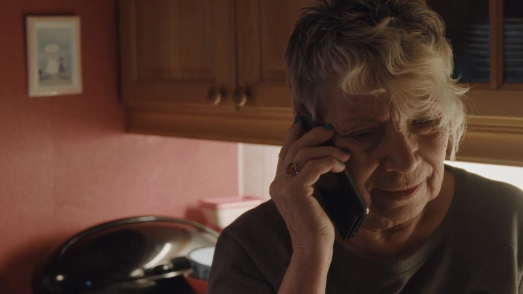 An older woman with short gray hair is talking on the phone in a kitchen with wooden cabinets and a pink wall.