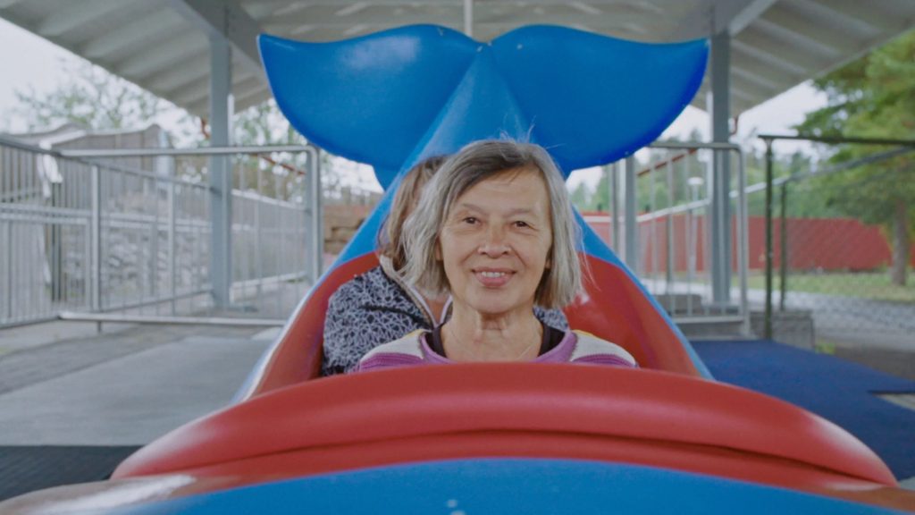 An older woman smiles while sitting in the front seat of a blue and red amusement park ride shaped like a whale. Another person sits behind her.