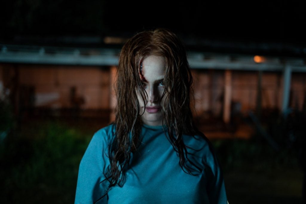 A woman with wet, disheveled hair and a serious expression stands outdoors at night, wearing a blue shirt, with a dimly lit building in the background.