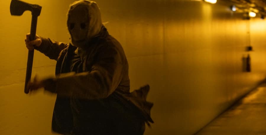 A person wearing a mask and holding an axe stands in a dimly lit, yellow-toned hallway.