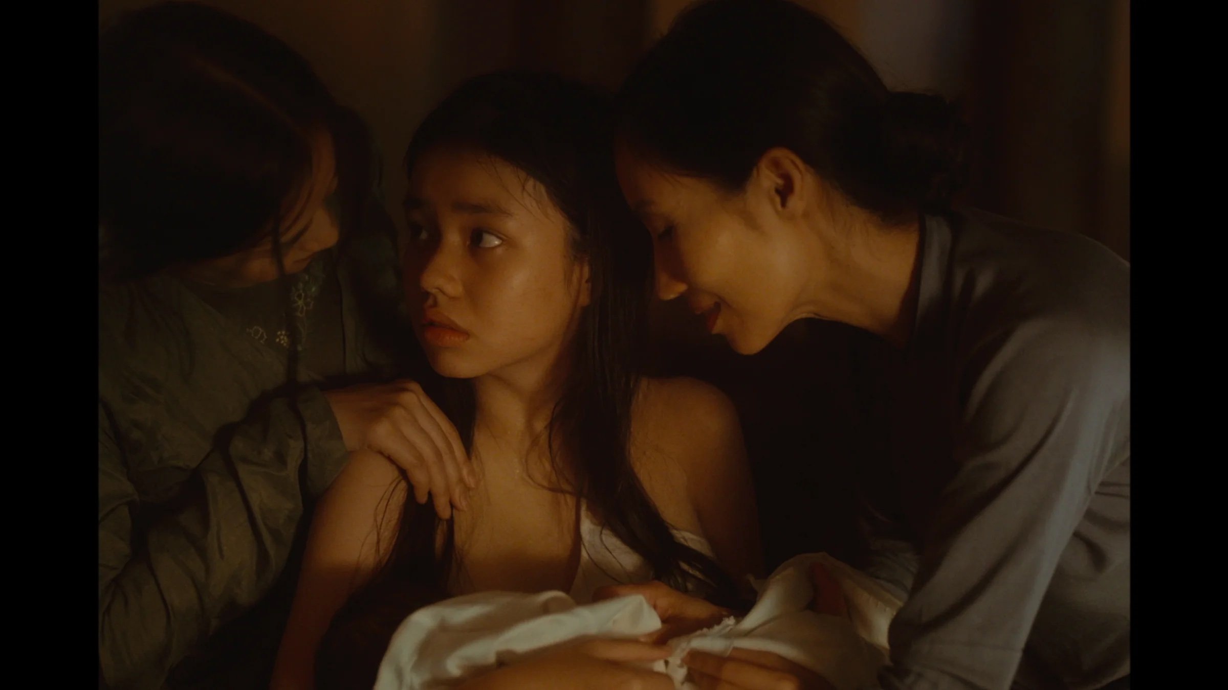 Three women sit close together in dim light; the central woman looks to the side while the others lean in, touching her supportively, and a white cloth is draped across their laps.