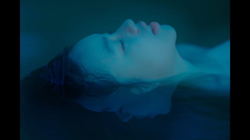 A person with closed eyes floats in water, their face partially submerged and reflected, with blue lighting creating a calm, surreal atmosphere.