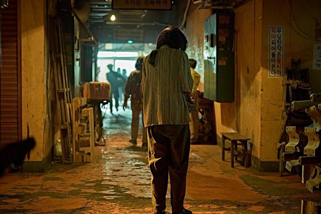 A person stands in a dimly lit, narrow hallway with other figures and various objects scattered along the walls; bright light shines from the far exit.