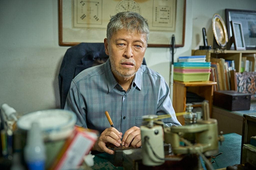 An older man with gray hair and beard sits at a cluttered desk, holding a tool and working on a small object, surrounded by various equipment and supplies.