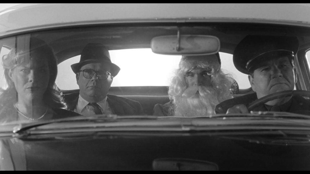 Four adults sit in a car, two in the front and two in the back; one passenger wears a fake white beard, and the image is in black and white.