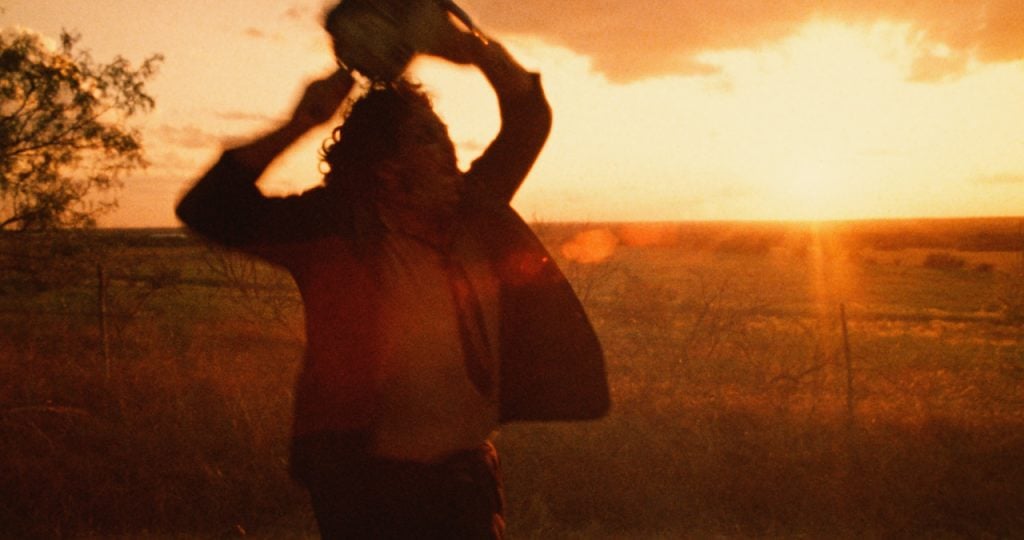 A person holding a chainsaw above their head stands in a field at sunset, silhouetted against the sky.
