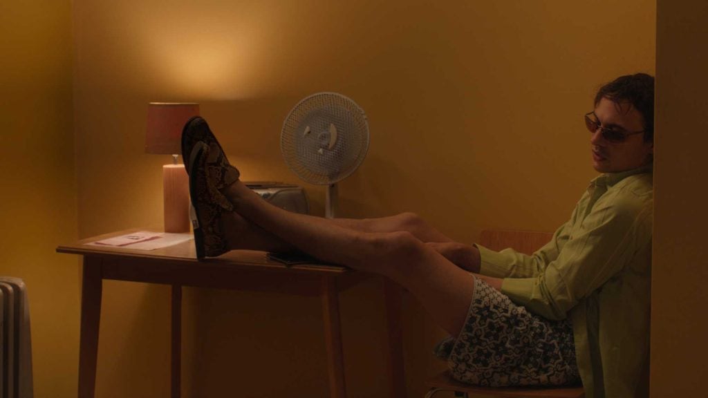 A man in sunglasses and casual clothes sits sideways on a chair with his feet on a desk in a dimly lit yellow room.
