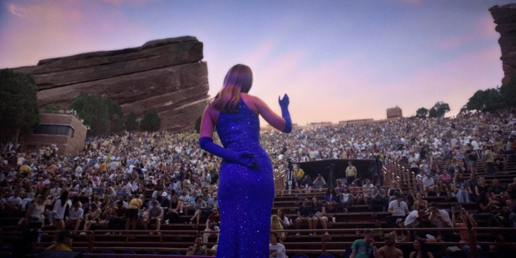 A performer in a sparkly blue dress and gloves stands on stage facing a large seated outdoor audience at sunset.