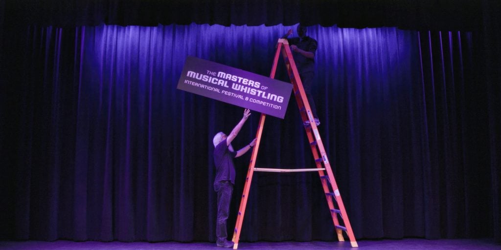 Two people set up a sign reading "The Masters of Musical Whistling International Festival & Competition" on stage in front of closed purple curtains.