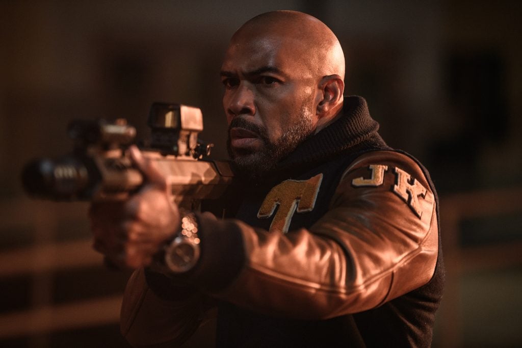 A man in a brown and black varsity jacket aims a rifle with a scope, appearing focused and intense in a dimly lit setting.