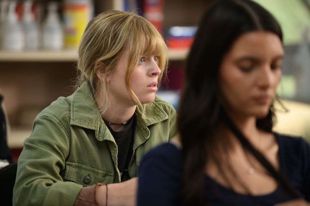 A young woman with blonde hair and a green jacket looks ahead attentively, while another woman with dark hair is blurred in the foreground.