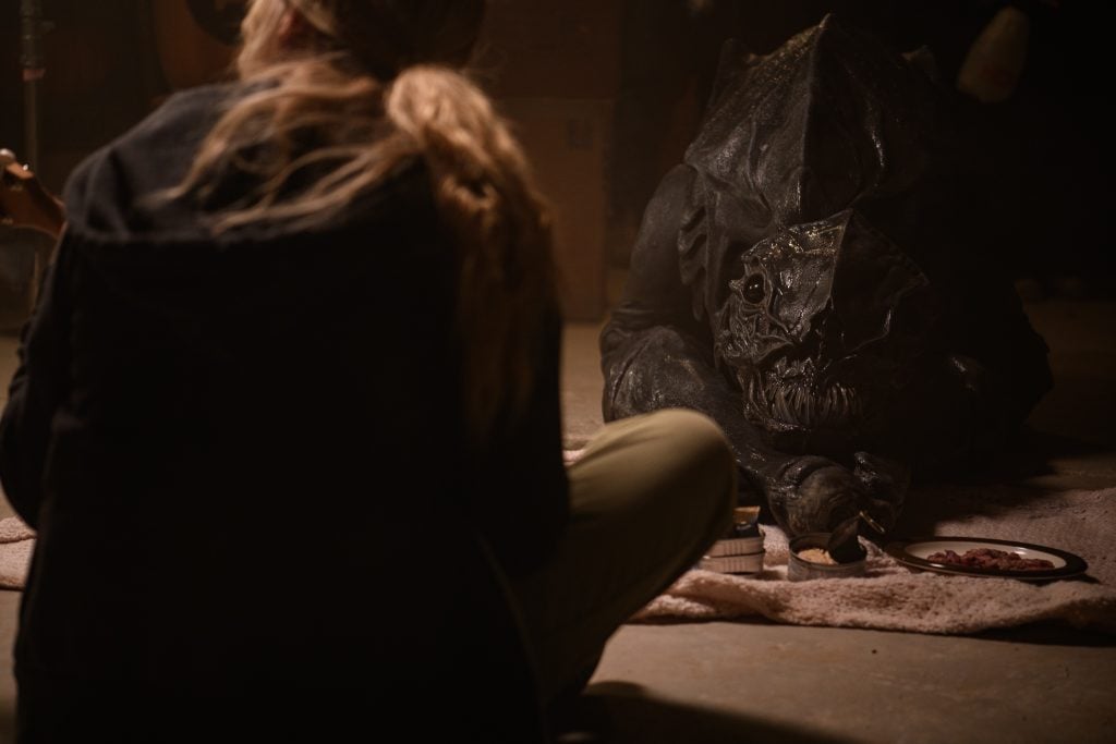 A person with light hair sits on the floor across from a creature in a dark, textured costume, both near objects and food placed on a blanket.