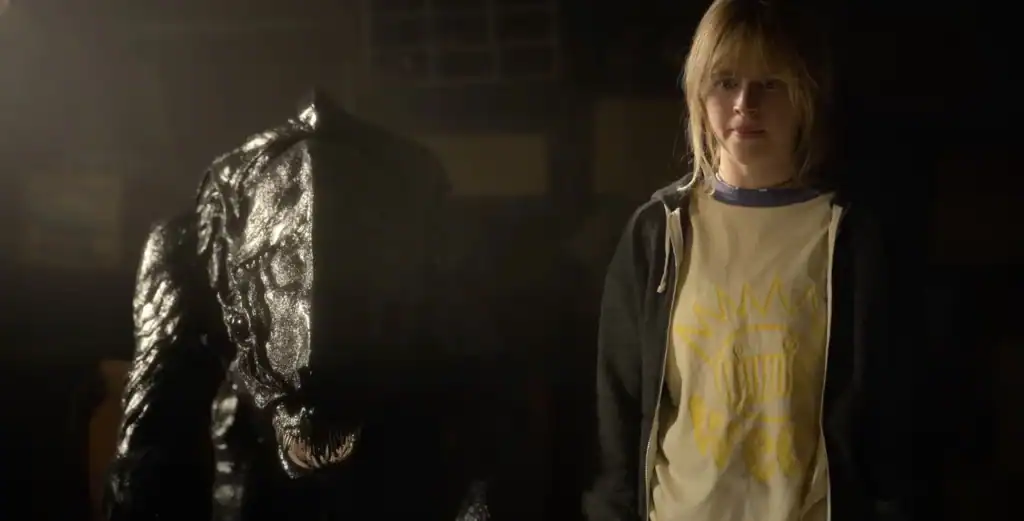 A young person with blonde hair stands next to a black, menacing, monster-like creature in a dimly lit room.