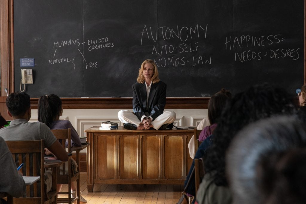 A woman sits cross-legged on a desk at the front of a classroom, speaking to students; notes on autonomy and happiness are written on the chalkboard behind her.