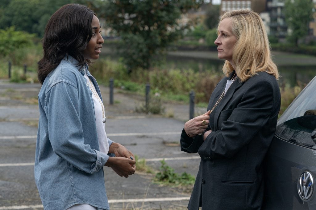 Two women stand and talk in an outdoor parking lot next to a car, with greenery and buildings visible in the background.