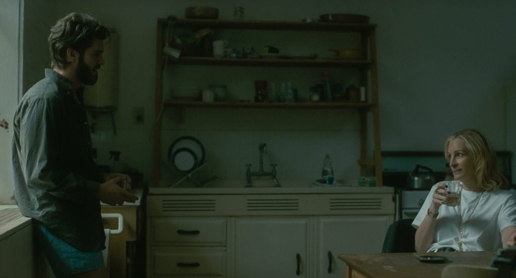 A man stands by a window in a dimly lit kitchen while a woman sits at a table holding a glass, with shelves and dishes visible in the background.