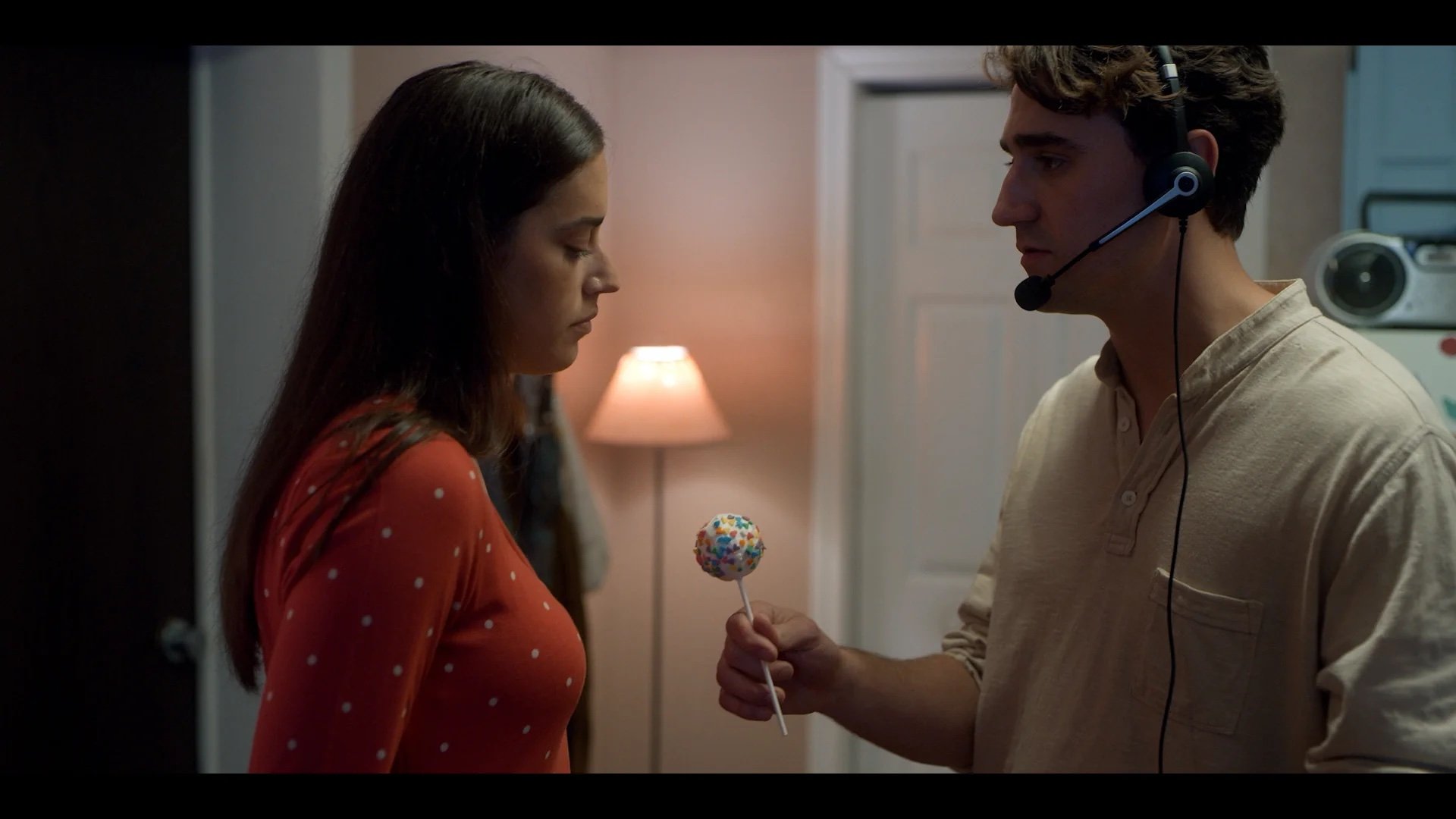 A man wearing a headset offers a cake pop with sprinkles to a woman in a polka dot shirt; both stand indoors near a lamp and door.