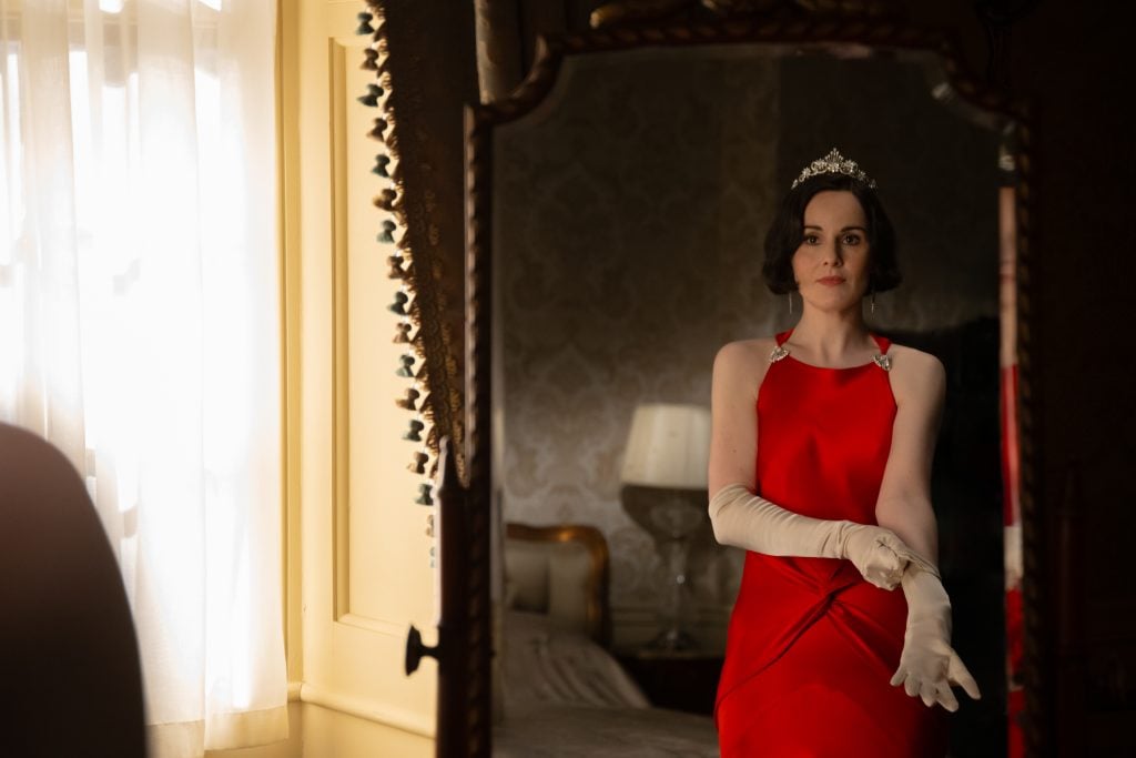 A woman in a red gown and tiara stands by a mirror, adjusting long white gloves in a softly lit room.