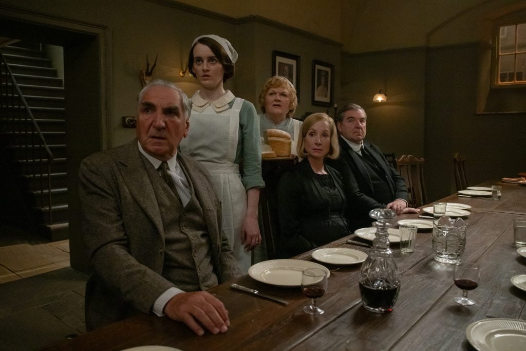 Five people, including a maid holding bread, sit at a long dining table set with plates, glasses, and a decanter, all looking intently in the same direction.