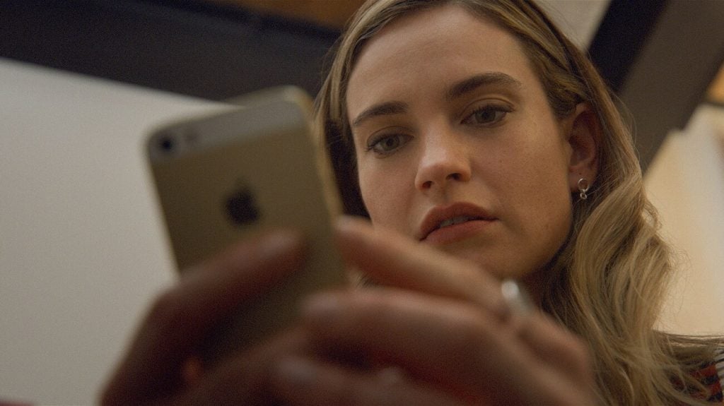 A woman with long hair looks intently at a smartphone she is holding in both hands, indoors under soft lighting.