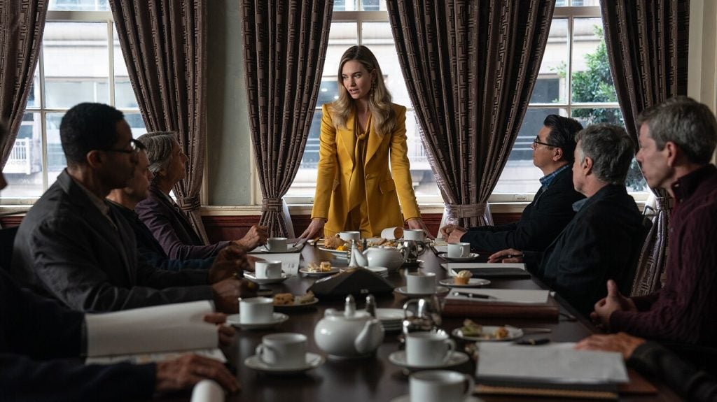 A woman in a yellow suit stands at a conference table, speaking to six seated individuals during a meeting in a room with large windows and curtains.