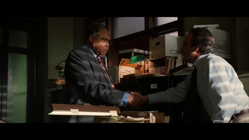 Two men shake hands in a cluttered office, surrounded by papers, folders, and file boxes.