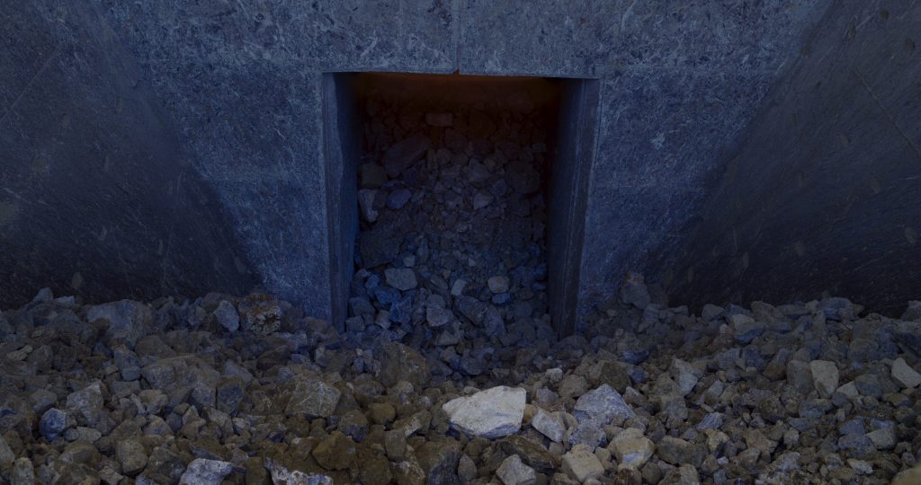A concrete chute or opening partially filled with loose rocks and rubble, with more rocks scattered on the ground in front.