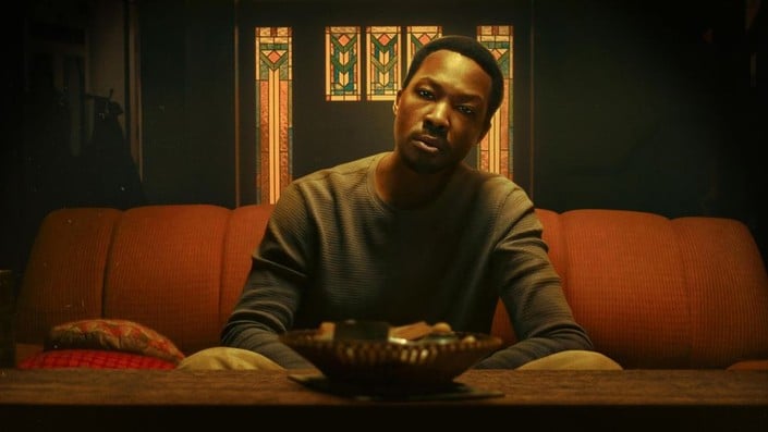 A man sits on an orange sofa in a dimly lit room, facing forward with a neutral expression. Stained glass panels are visible in the background.