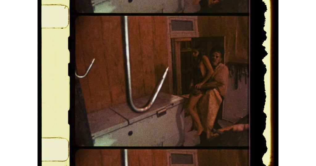 A film still shows two people wrapped in towels near a chest freezer in a dimly lit room with wood-paneled walls and a large metal hook in the foreground.