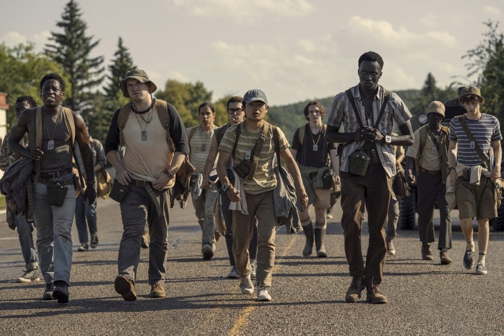 A group of people in casual outdoor clothing and backpacks walk down a paved road, appearing determined and organized, with trees and blue sky in the background.