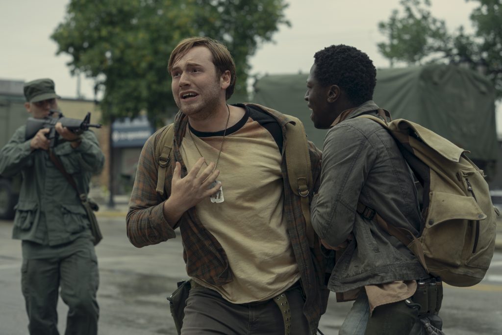 Two men with backpacks run in distress on a street while a soldier aims a rifle at them from behind; military vehicles and trees are visible in the background.