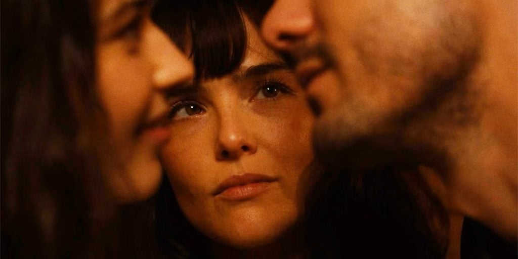 A woman looks intently at two people in the foreground, whose faces are out of focus, suggesting a moment of contemplation or observation.