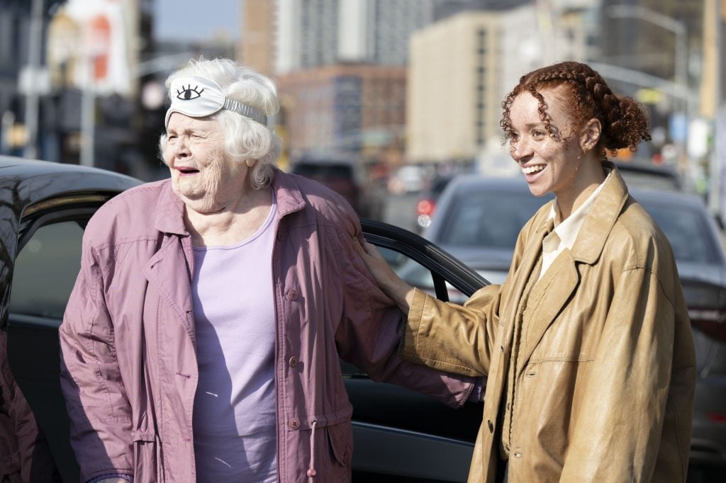 An elderly woman wearing a sleep mask on her head smiles as she stands next to a younger woman by an open car door on a city street.