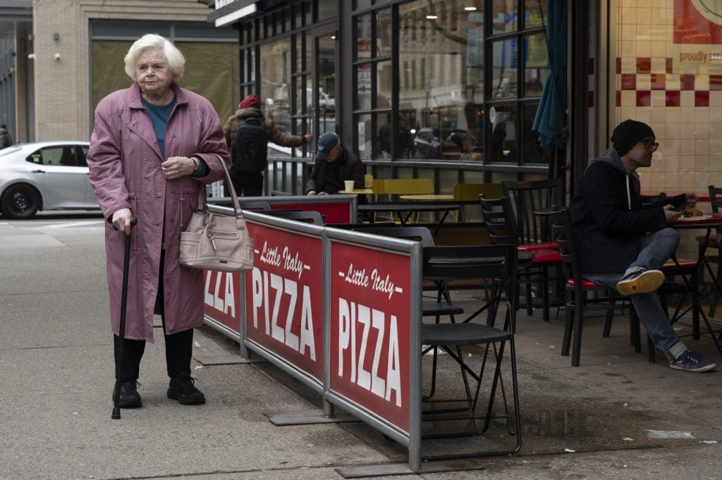 An elderly woman in a pink coat and holding a cane stands on the sidewalk outside a pizza restaurant; people sit at tables nearby.