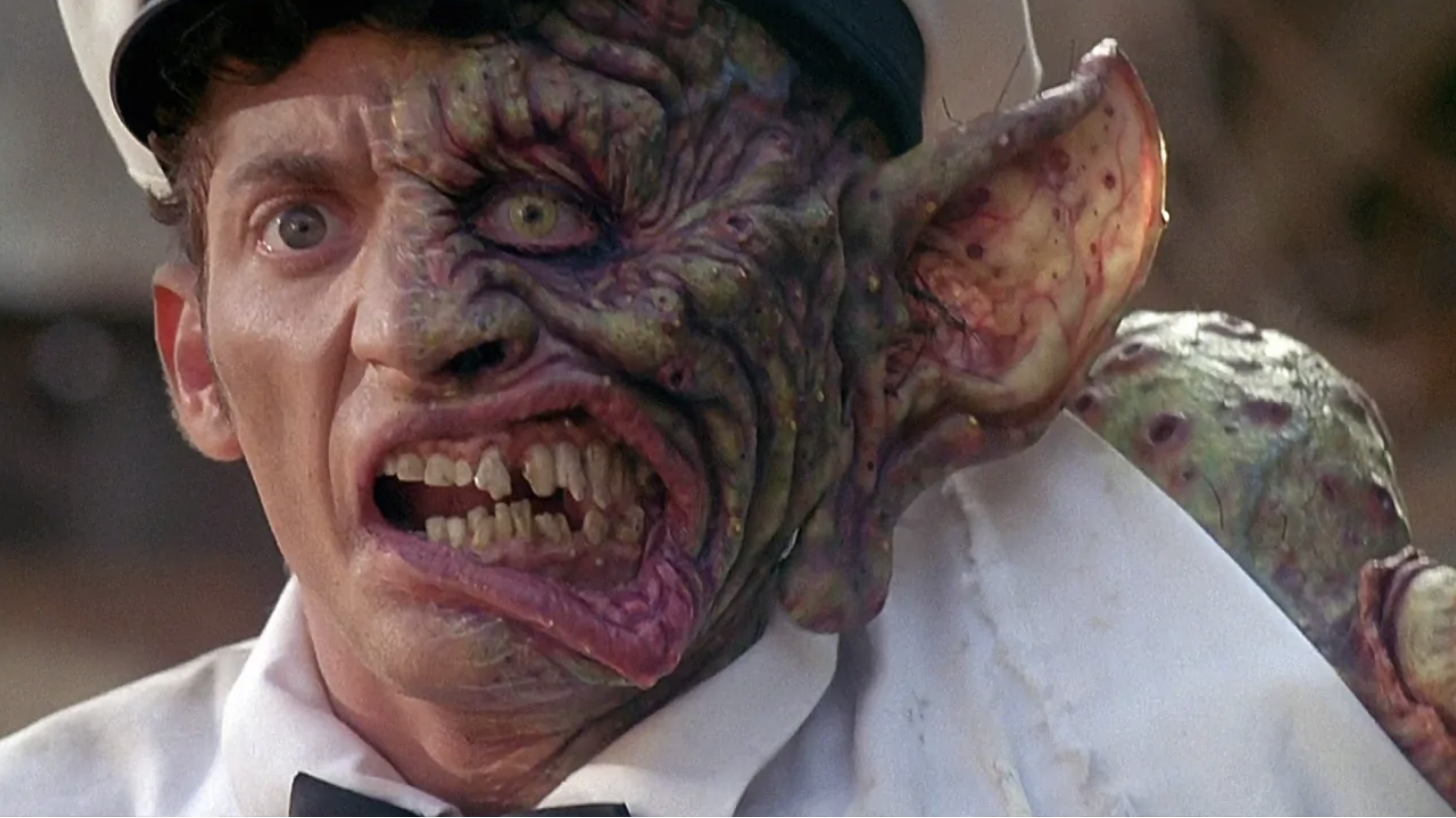 A man in a white uniform has one half of his face transformed into a grotesque, monstrous shape with discolored skin, sharp teeth, and a large, deformed ear.