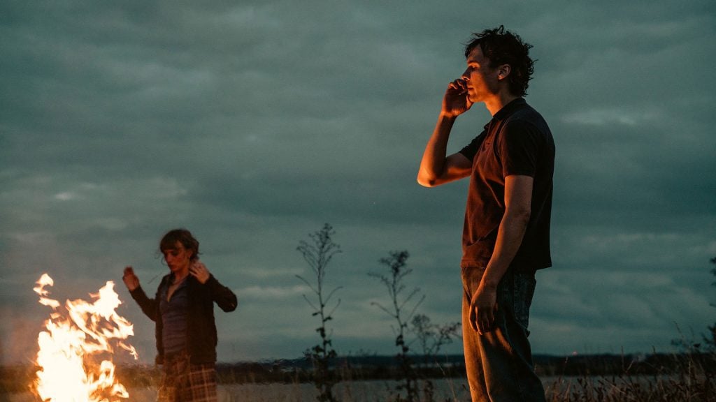A person stands near a small fire at dusk, talking on the phone, while another person raises their arms beside the flames under a cloudy sky.