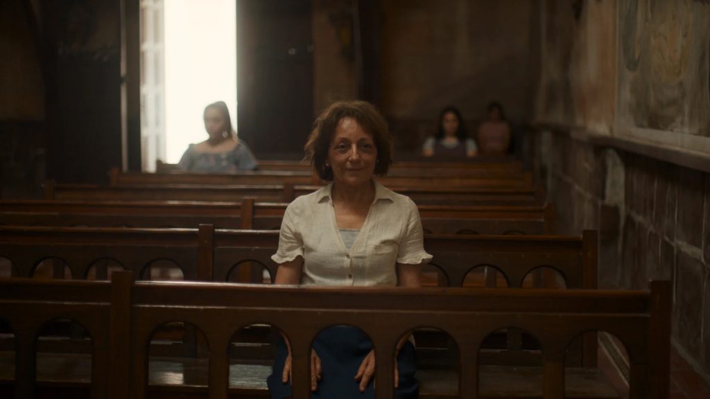 A woman sits alone on a wooden pew in a dimly lit church, with three other people seated separately in the background. Light enters through an open door behind her.