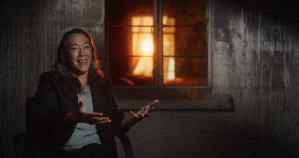 A woman sits in a dimly lit room, gesturing with her hands while talking. A window behind her shows warm light shining through.
